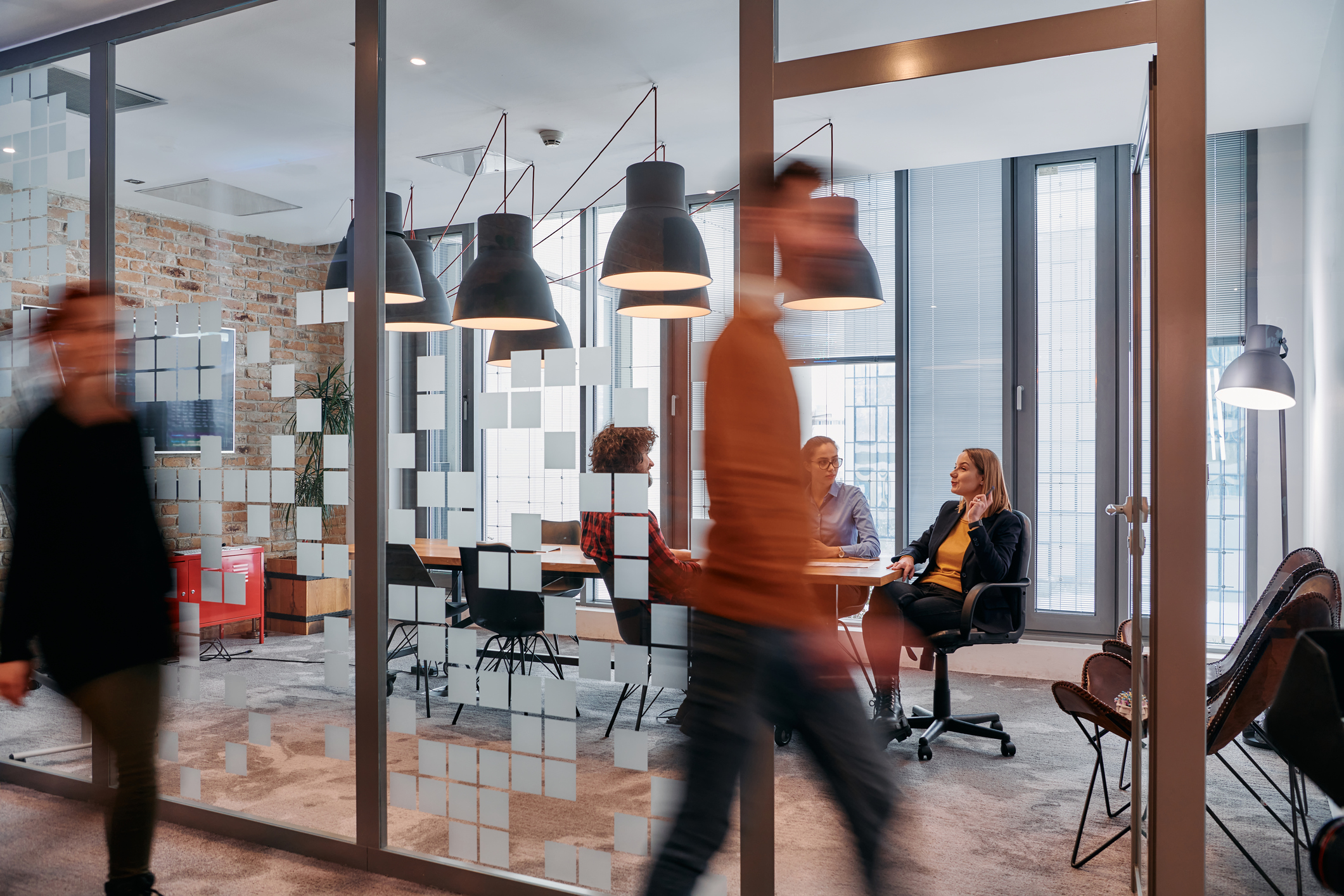 In the dynamic hustle and bustle of a business environment, a group of young business professionals walk down the corridor next to their office, while their colleagues collaborate inside, symbolizing teamwork and productivity